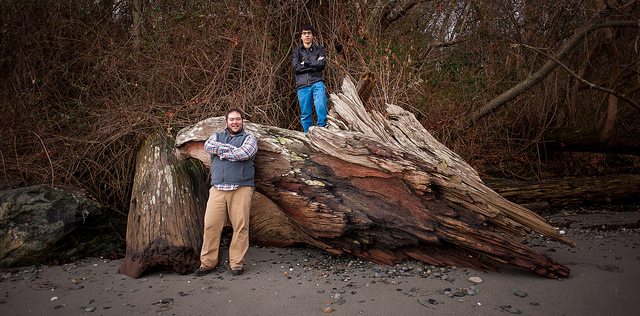 Two men pose against an old fallen tree on a beach.